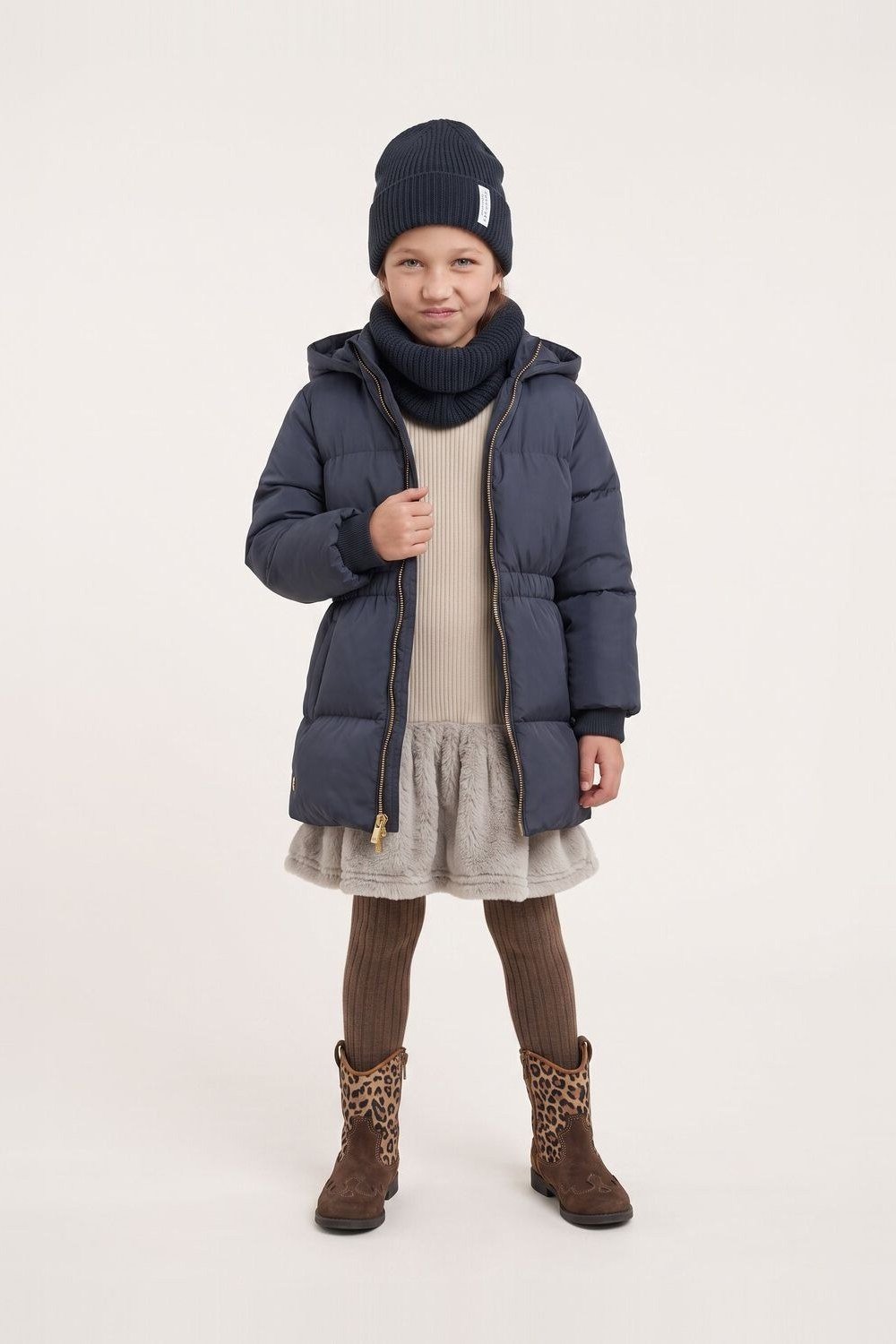 Woman over 60 wearing a navy puffer coat over a cream dress with brown boots and beanie