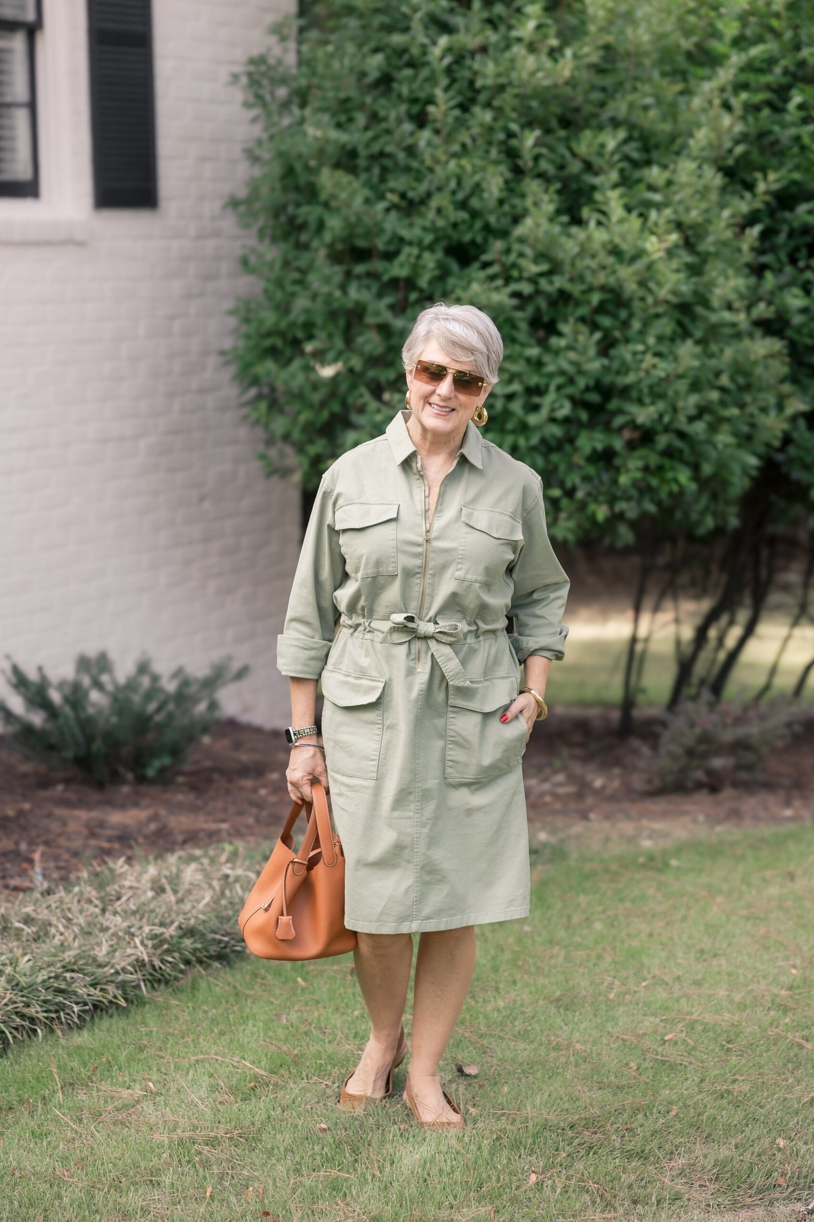 Sage safari shirt dress for women over 60: tie belt and flap pockets with a tan bag in a garden setting