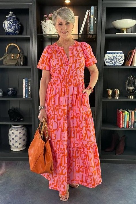 Older woman wearing coral red patterned maxi dress, bookshelf interior, a timeless casual outfit for women over 60.