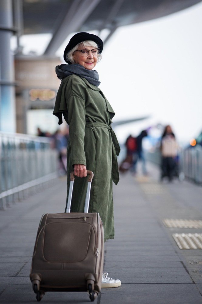 Older woman wearing olive green long coat, dark rolling suitcase, airport platform, a timeless casual outfit for women over 60.