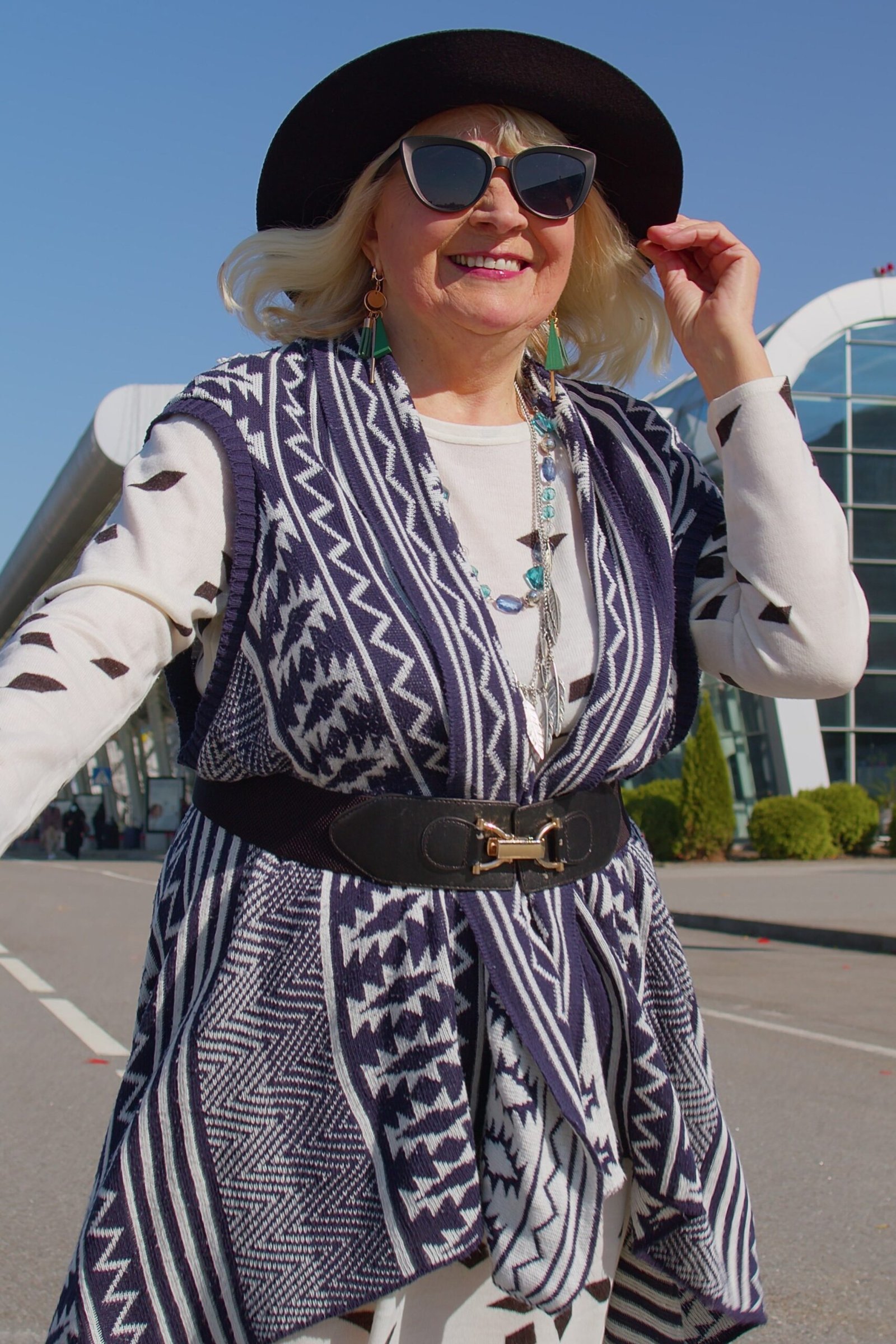 Older woman wearing blue patterned vest and white top and hat, airport curb, a timeless casual outfit for women over 60.