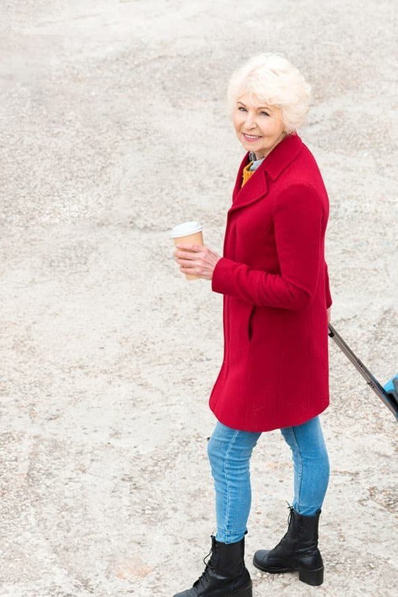Older woman wearing red wool coat and blue jeans, rolling blue suitcase, aerial view, a timeless casual outfit for women over 60.