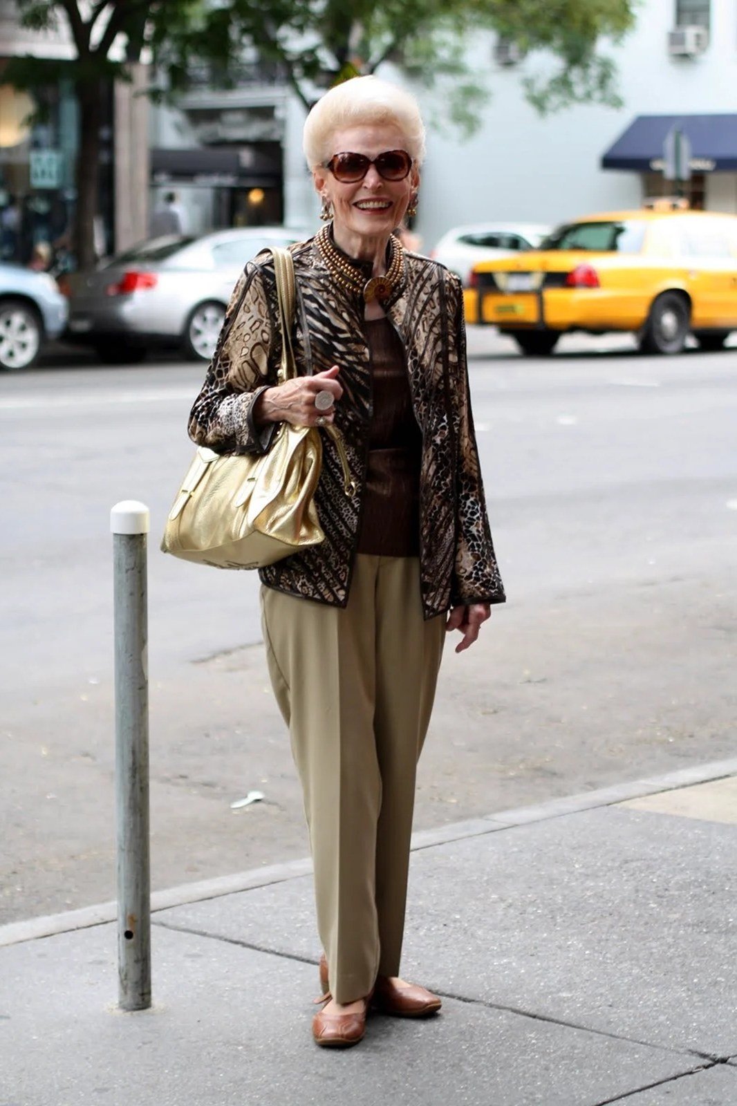 Older woman wearing brown patterned jacket and khaki wide pants, city sidewalk, a timeless casual outfit for women over 60.