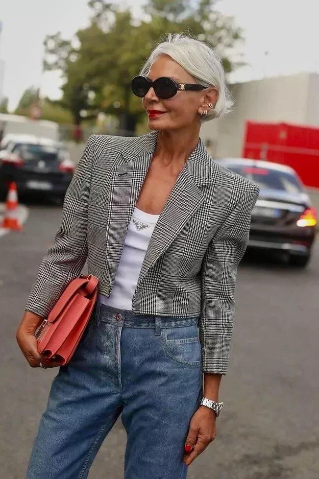 Older woman wearing gray cropped blazer and white tee and blue jeans and red bag, street, a timeless casual outfit for women over 60.