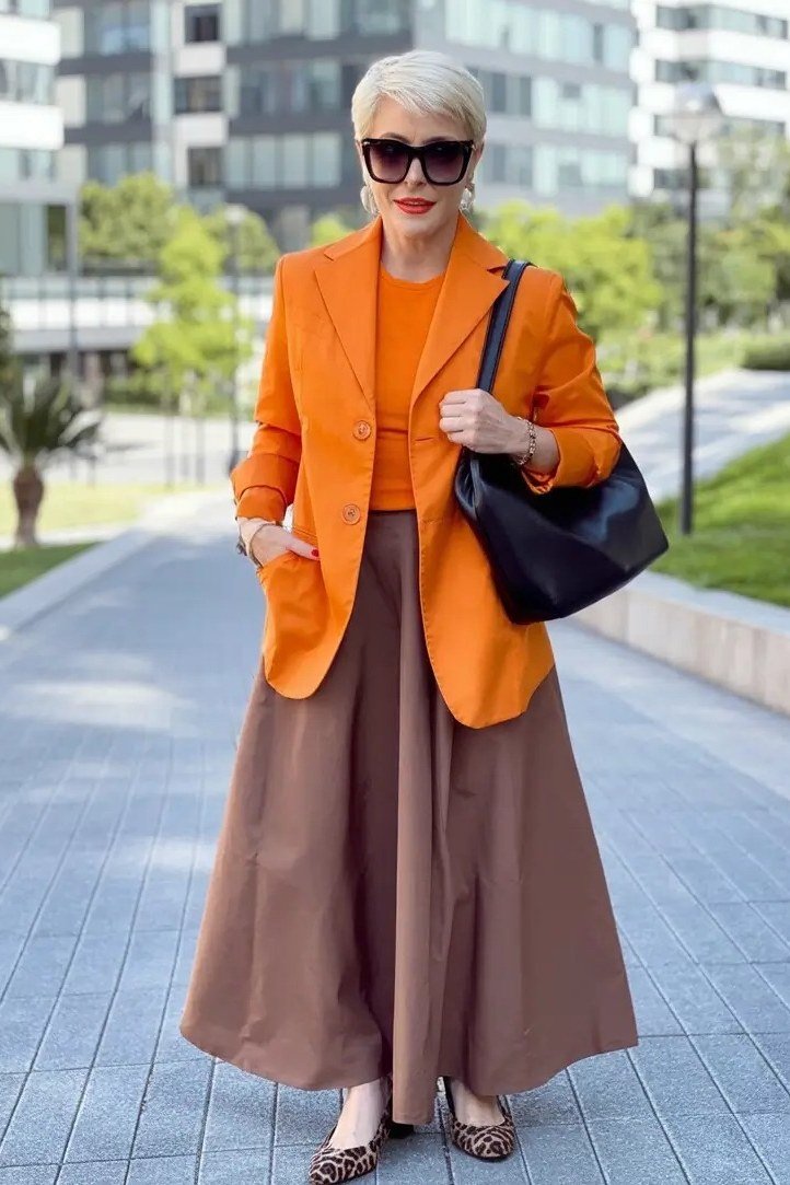 Older woman wearing burnt orange blazer and chocolate brown midi skirt, city, a timeless casual outfit for women over 60.