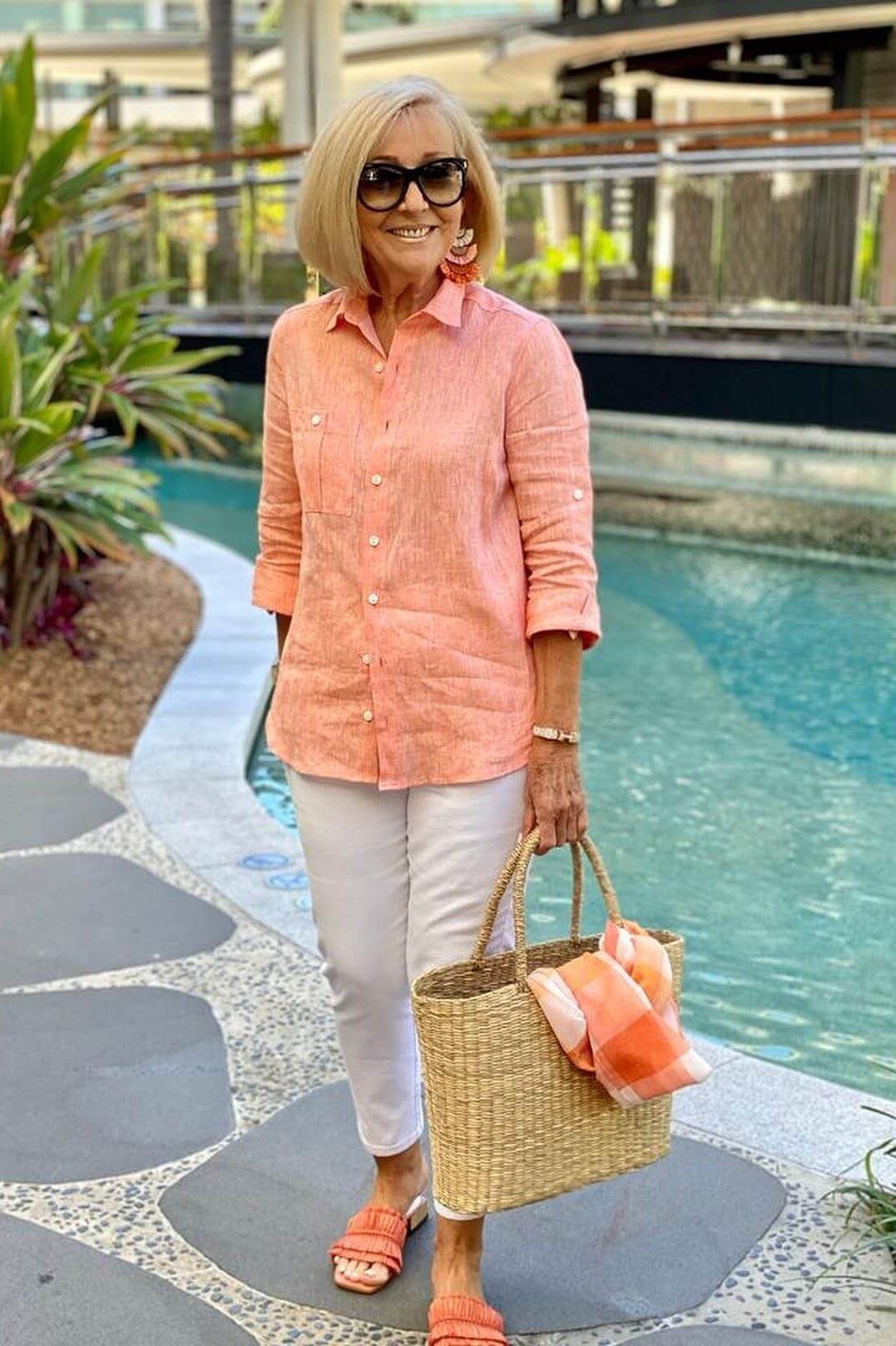 Older woman wearing coral linen shirt and white capris and straw tote, poolside, a timeless casual outfit for women over 60.