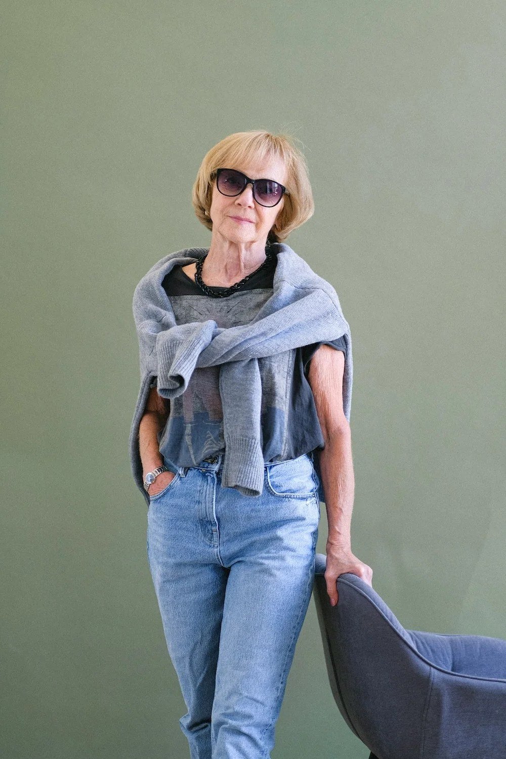 Older woman wearing gray knit sweater and light wash jeans, sage wall, a timeless casual outfit for women over 60.