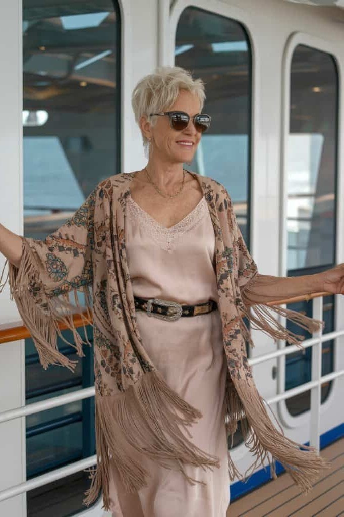 Beige tank top with a blush pink satin midi skirt, thin dark belt, and a fringed taupe kimono, on a cruise ship deck; soft romantic cruise layers - travel outfit for women over 50