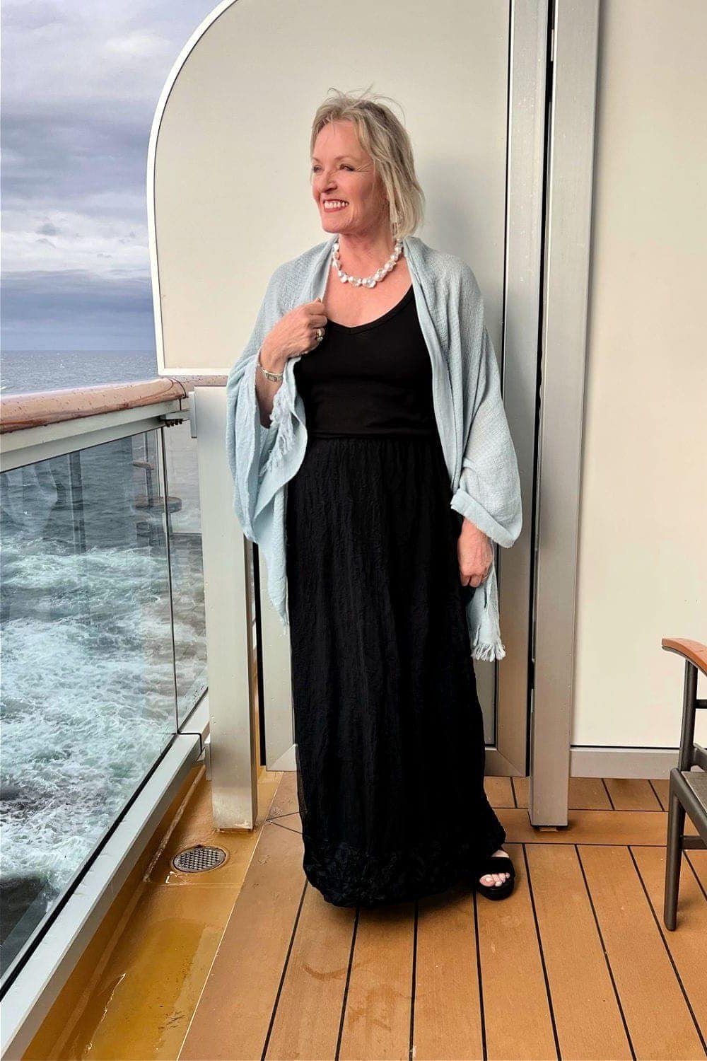 Black maxi dress with a light blue open cardigan draped over the shoulders, on a cruise ship balcony; simple chic evening cruise look - travel outfit for women over 50