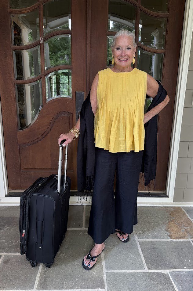 Mustard yellow sleeveless top with black wide-leg pants and black sandals, standing with a rolling suitcase; cheerful high-contrast travel day outfit - travel outfit for women over 50