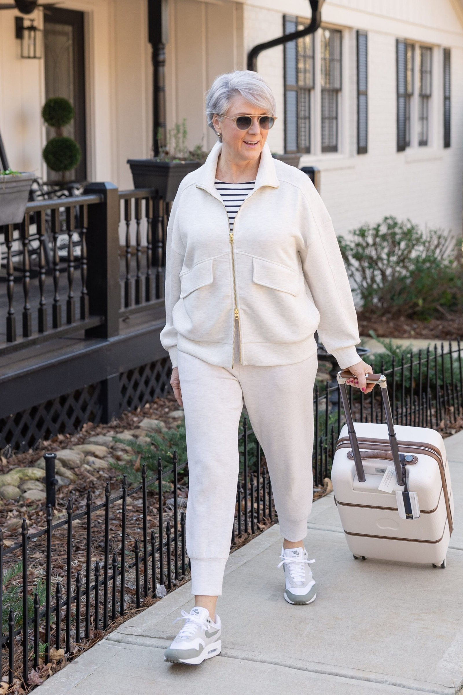 All-white outfit of a relaxed button shirt and white pants with white sneakers, rolling a cream suitcase on a porch; fresh minimalist airport look - travel outfit for women over 50