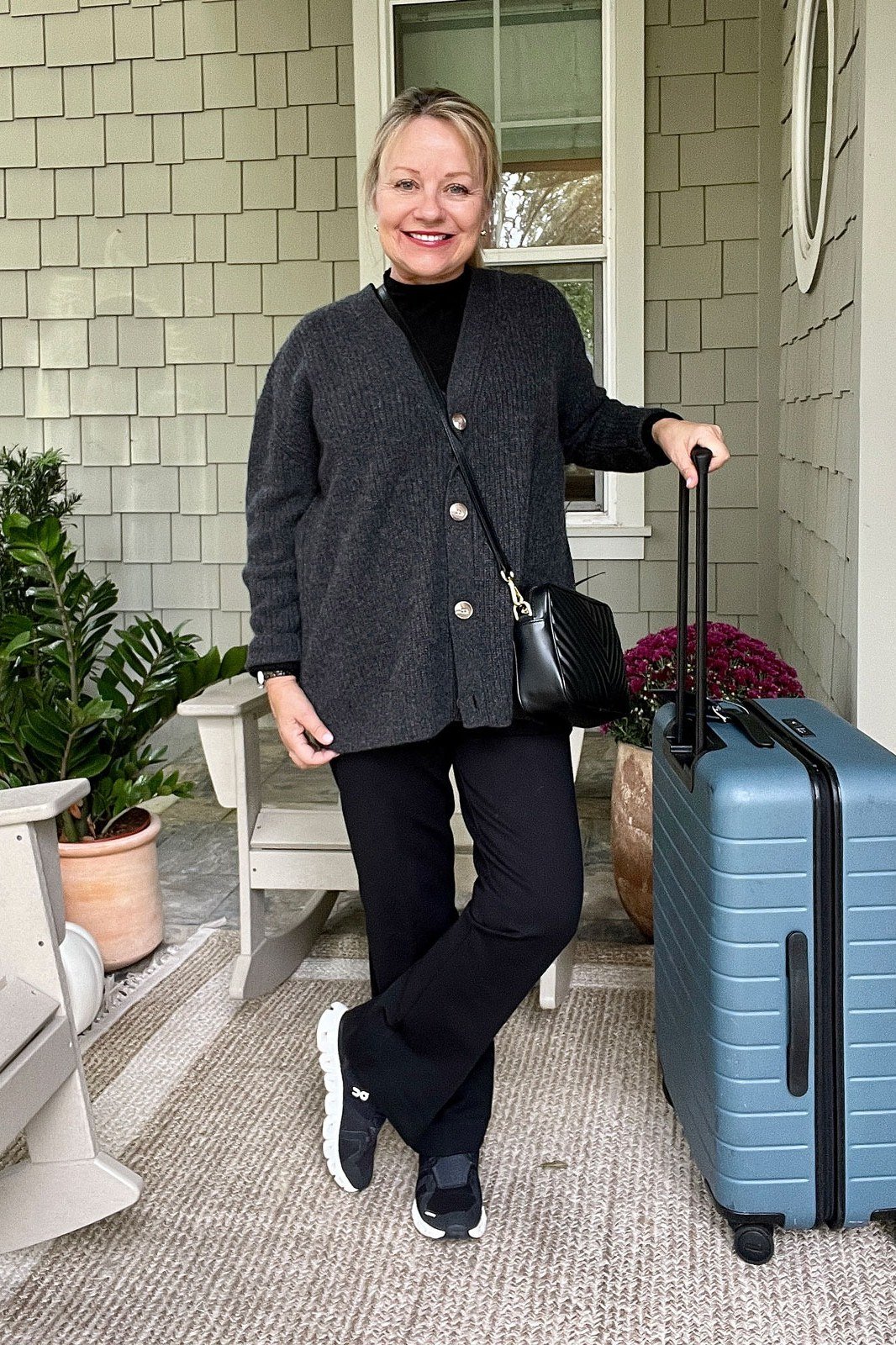 All-black outfit of a loose top and pants with black sneakers, leaning on a light blue suitcase on a porch; sleek monochrome travel uniform - travel outfit for women over 50