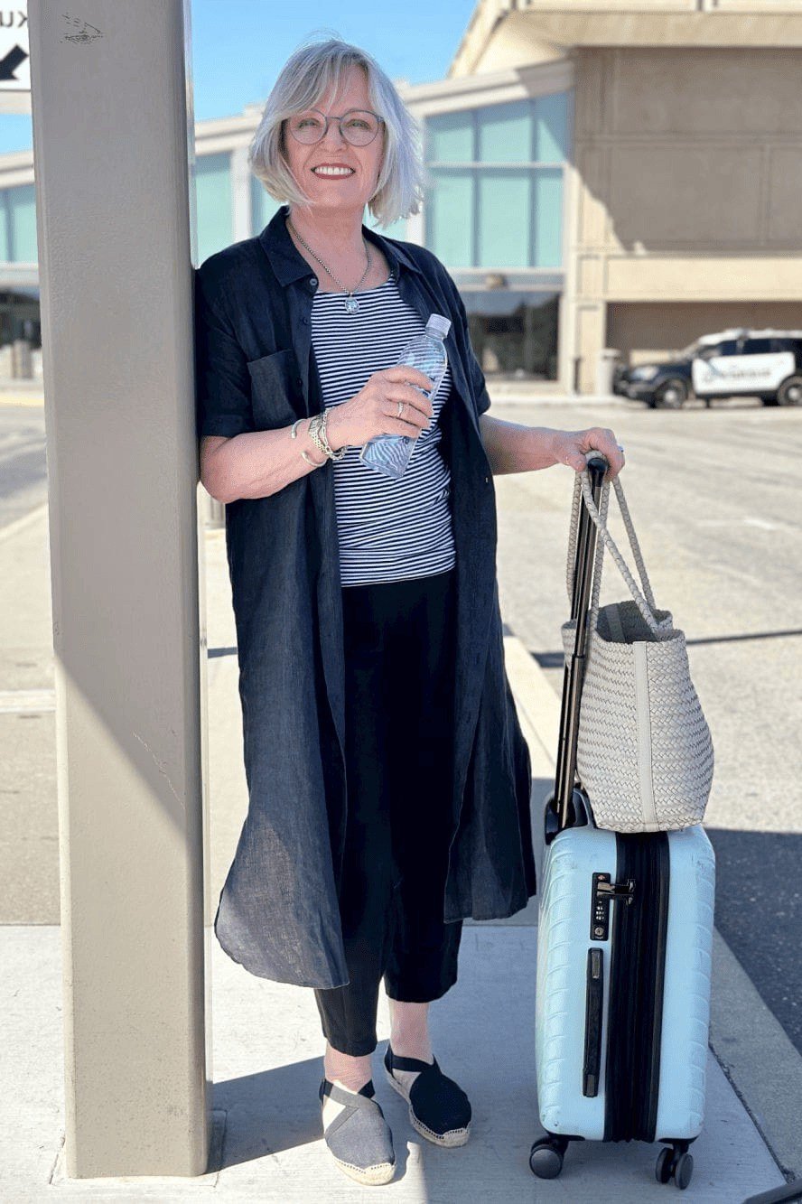 Navy long duster coat over a gray top and black pants, with a woven tote and rolling suitcase at an airport; layered airport travel outfit - travel outfit for women over 50