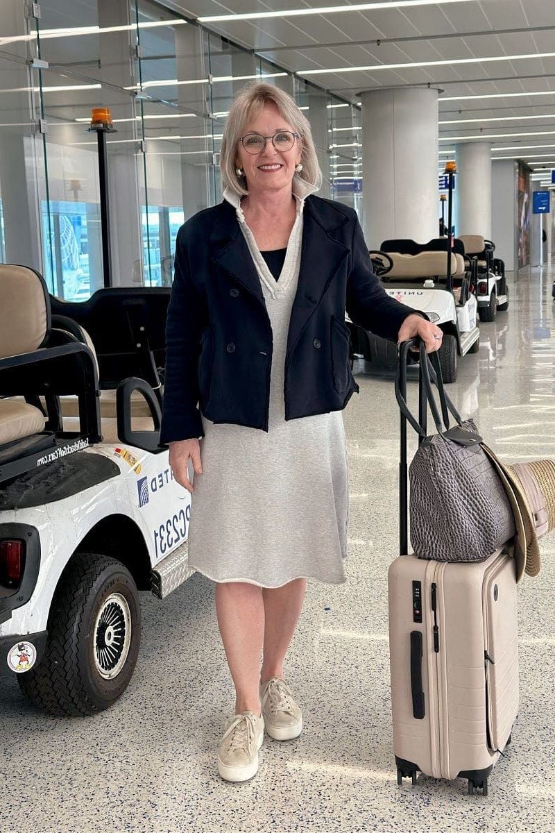 Navy blazer over a white midi dress with nude sneakers, rolling luggage beside an airport cart; smart casual airport outfit - travel outfit for women over 50