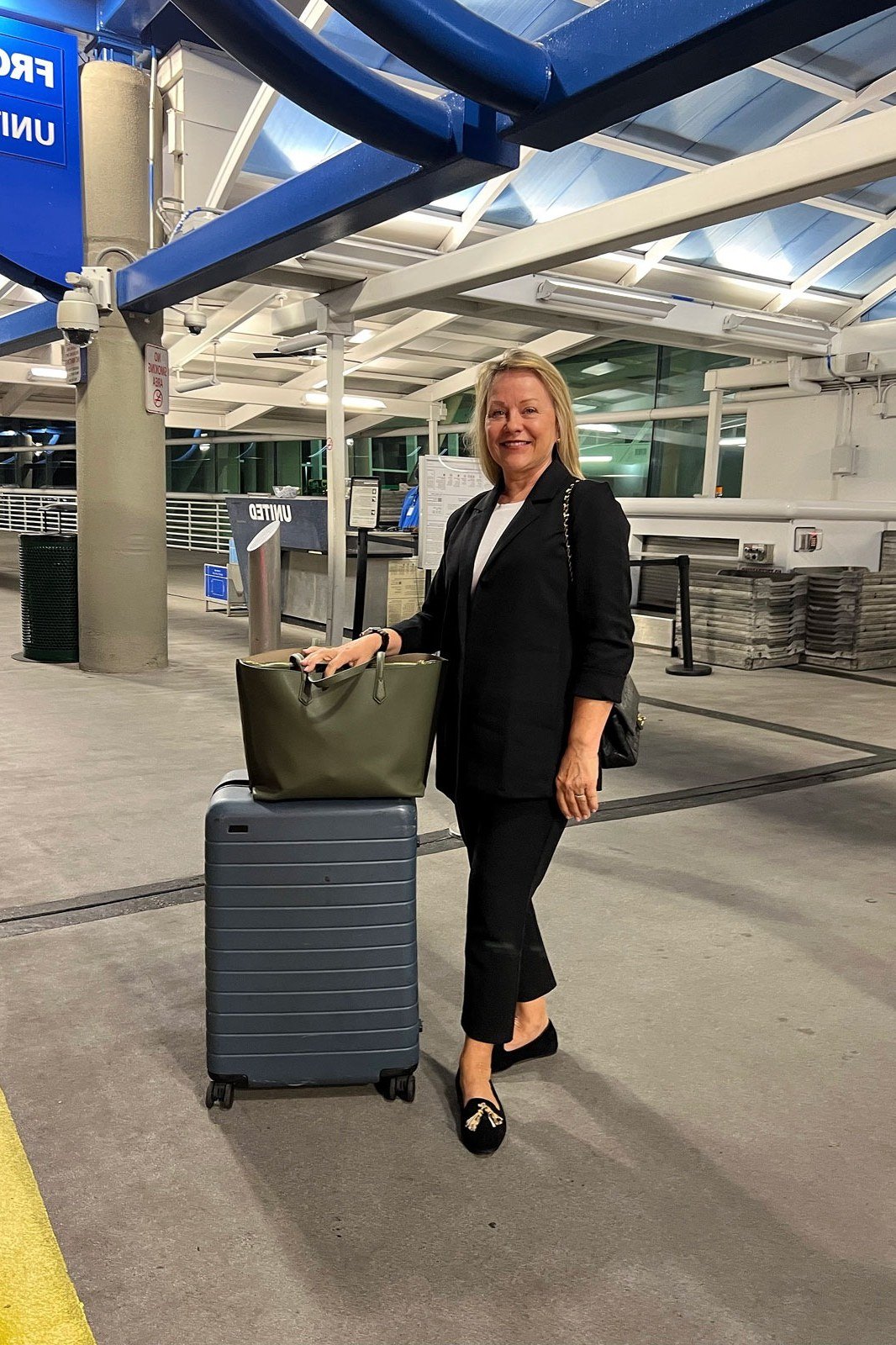 Black blazer with a black top and black pants, pulling a gray suitcase through an airport terminal; sharp all-black business travel look - travel outfit for women over 50