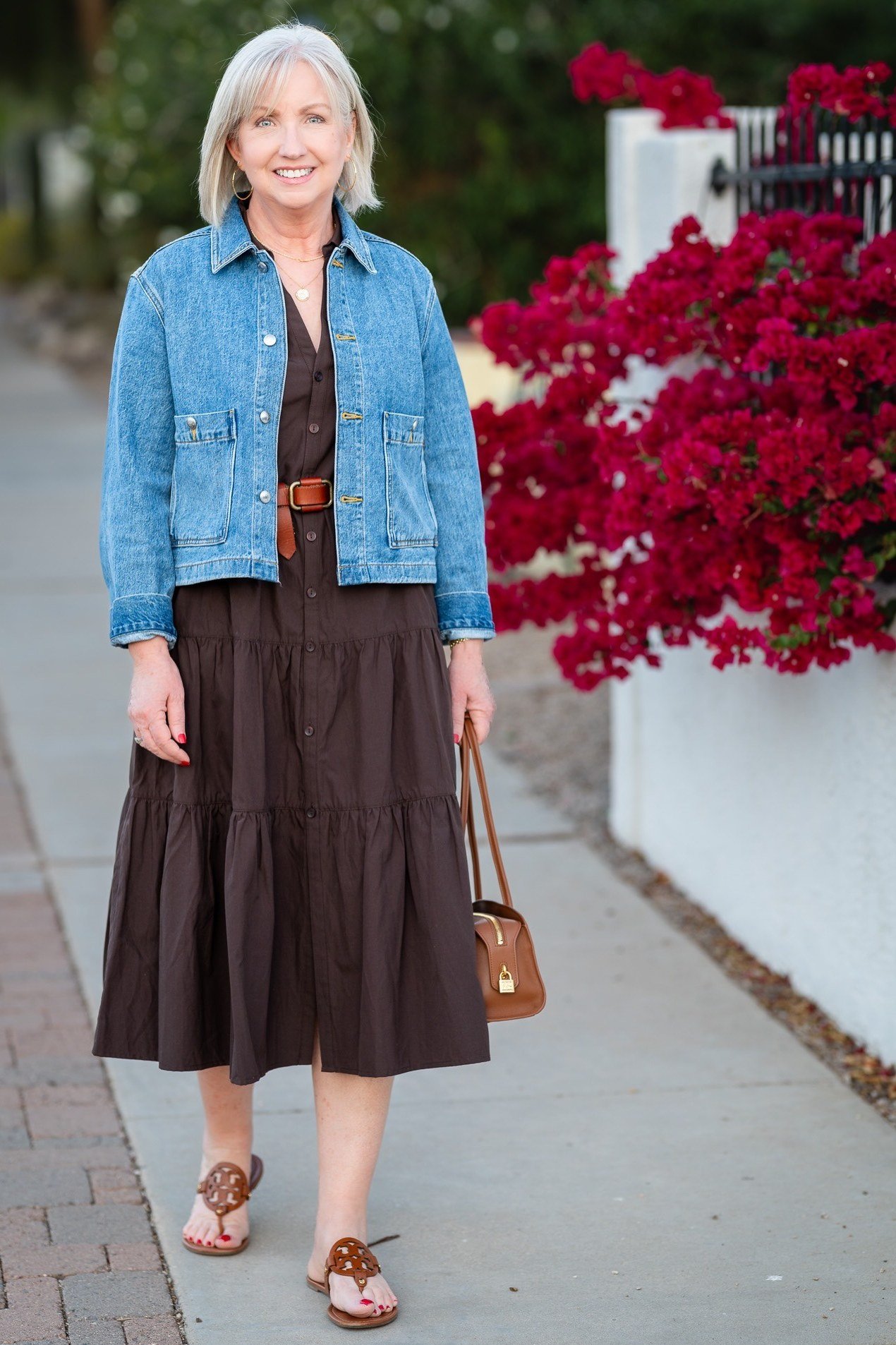 Light-wash denim jacket over a dark brown tiered maxi skirt with a tan shoulder bag and brown sandals, on a city street; denim-and-maxi street style - travel outfit for women over 50