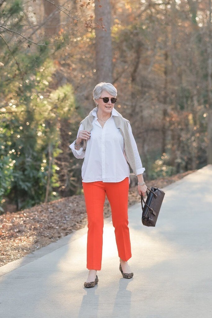 White button shirt with bright red cropped pants, dark sunglasses, and a small clutch, walking a park path; bold red-pants fall street look - travel outfit for women over 50