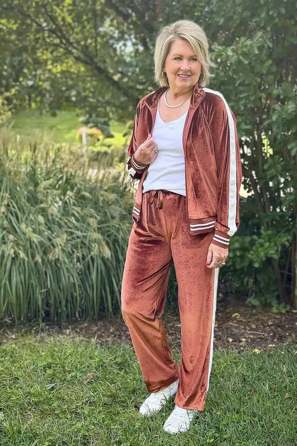 Rust-colored velour tracksuit with a white tee and white sneakers, standing in a garden; cozy retro travel tracksuit - travel outfit for women over 50