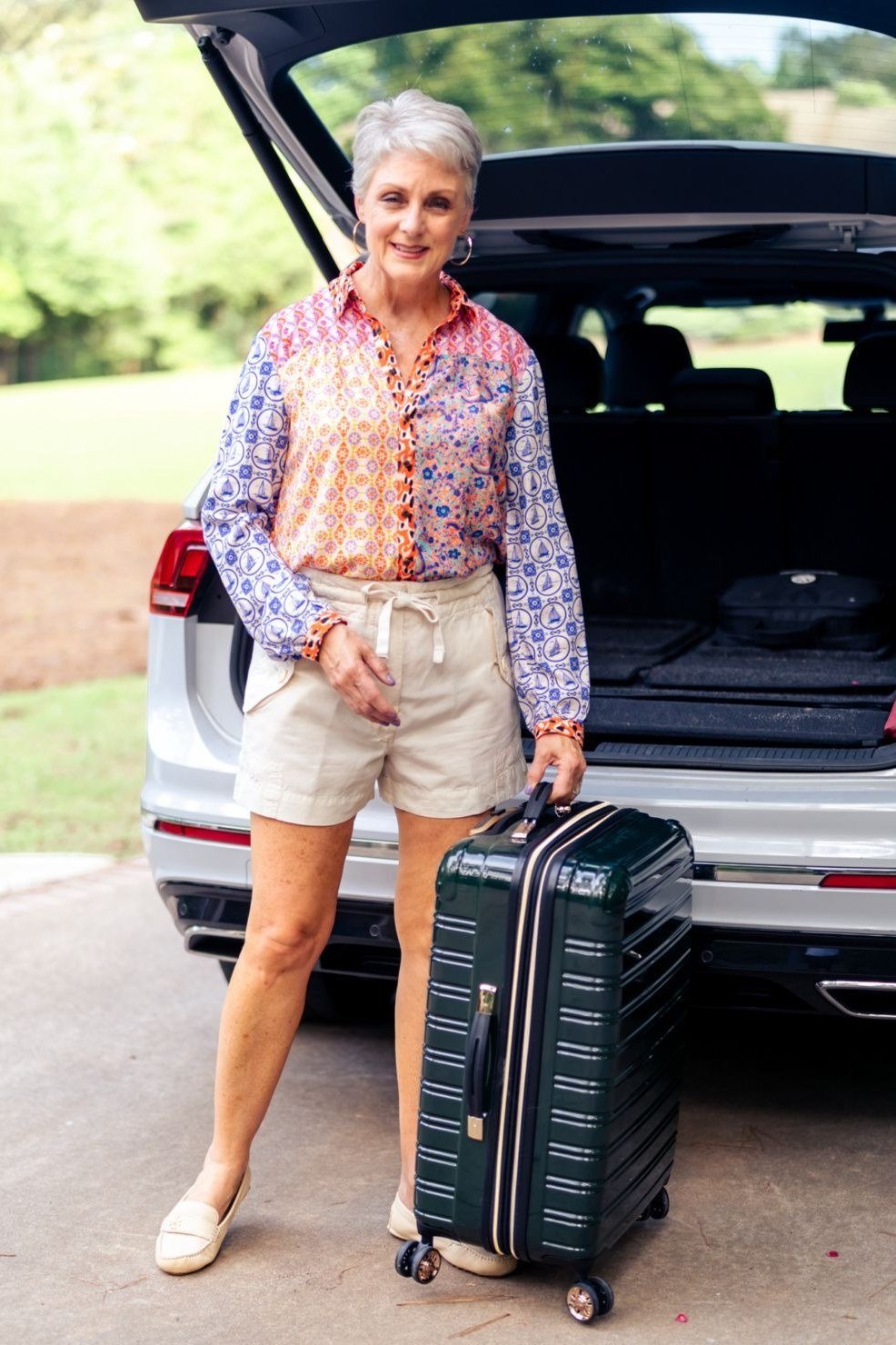 Pink and blue floral blouse with beige tailored shorts and nude flats, loading a suitcase at a car trunk; cheerful road trip outfit - travel outfit for women over 50