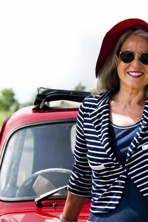 Navy-and-white striped top with a red sun hat and sunglasses, leaning on a small red car on a country road; fun retro road trip look - travel outfit for women over 50