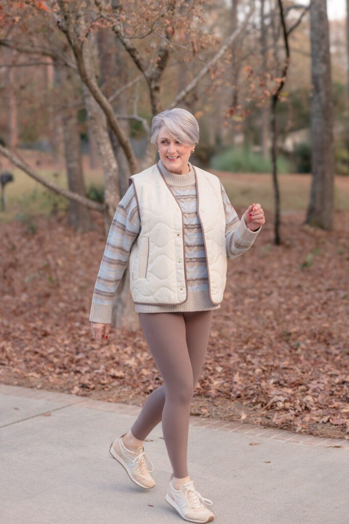 Fall turtleneck outfit for women over 50: striped mock-neck sweater with cream quilted vest, taupe leggings, autumn path