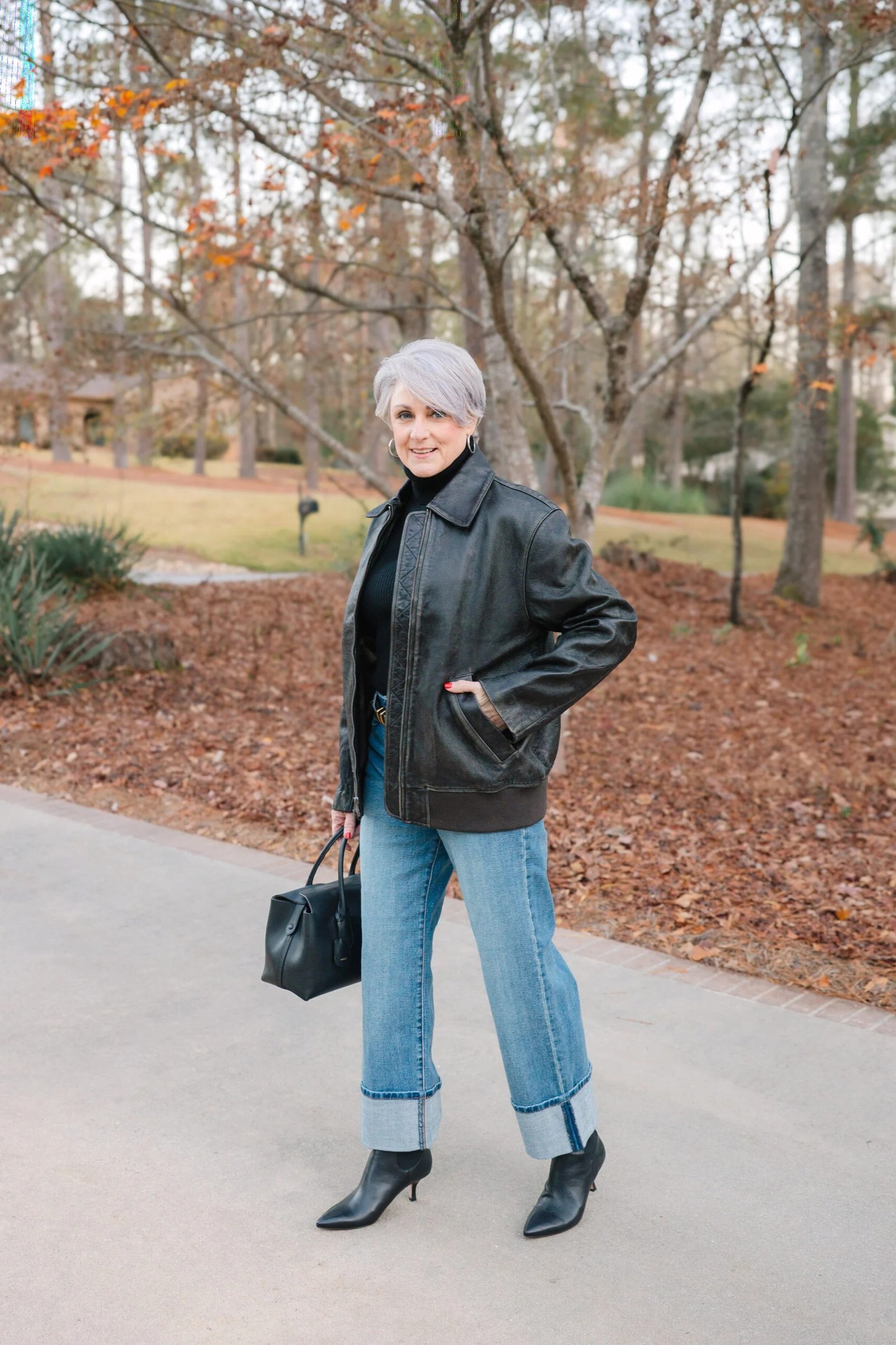 Fall turtleneck outfit for women over 50: black turtleneck with black leather bomber, cuffed jeans, black boots, autumn path