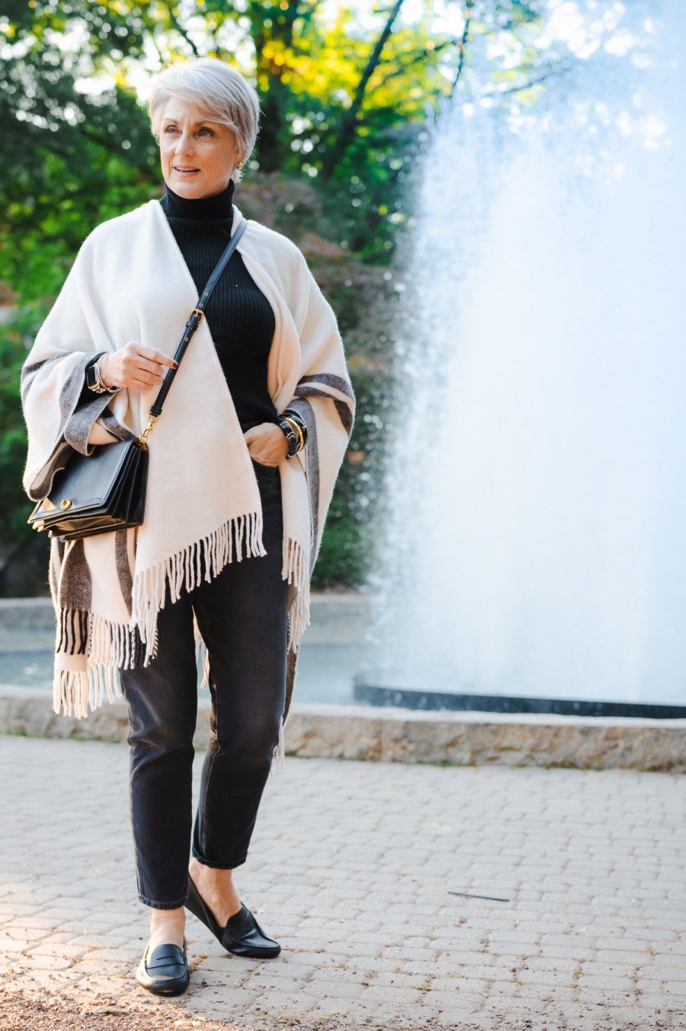 Fall turtleneck outfit for women over 50: black turtleneck with cream striped fringe poncho, fountain