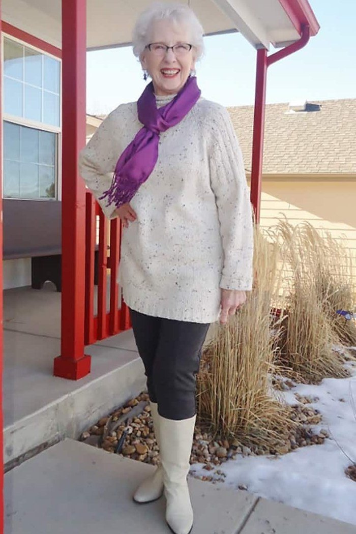 Older woman in cream cable knit sweater with purple scarf, dark jeans and cream ankle boots