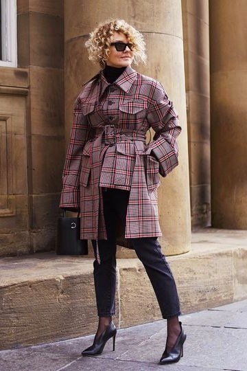 Older woman wearing mauve plaid belted trench coat with black cropped trousers and black heels