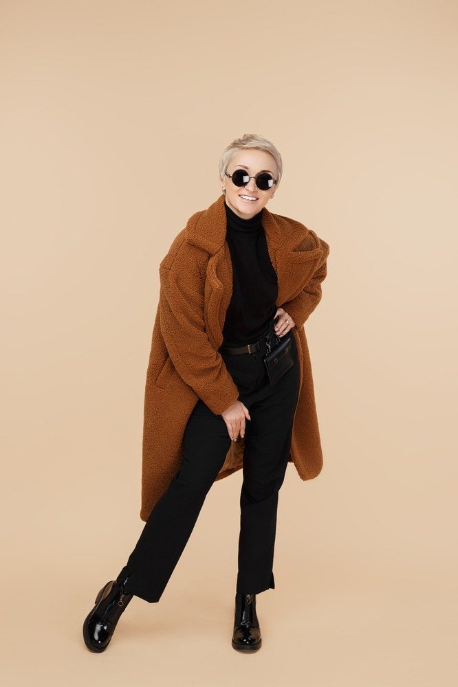 Older woman wearing rust brown wrap coat with black turtleneck and black cropped jeans