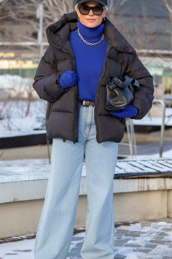 Woman wearing black puffer vest over royal blue turtleneck with light wash jeans and black cap