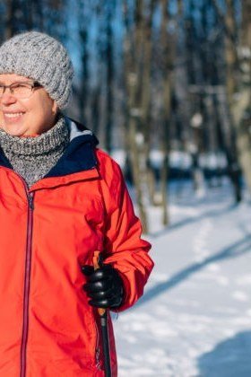 Older woman in red ski jacket with grey knit beanie holding ski poles in snowy forest