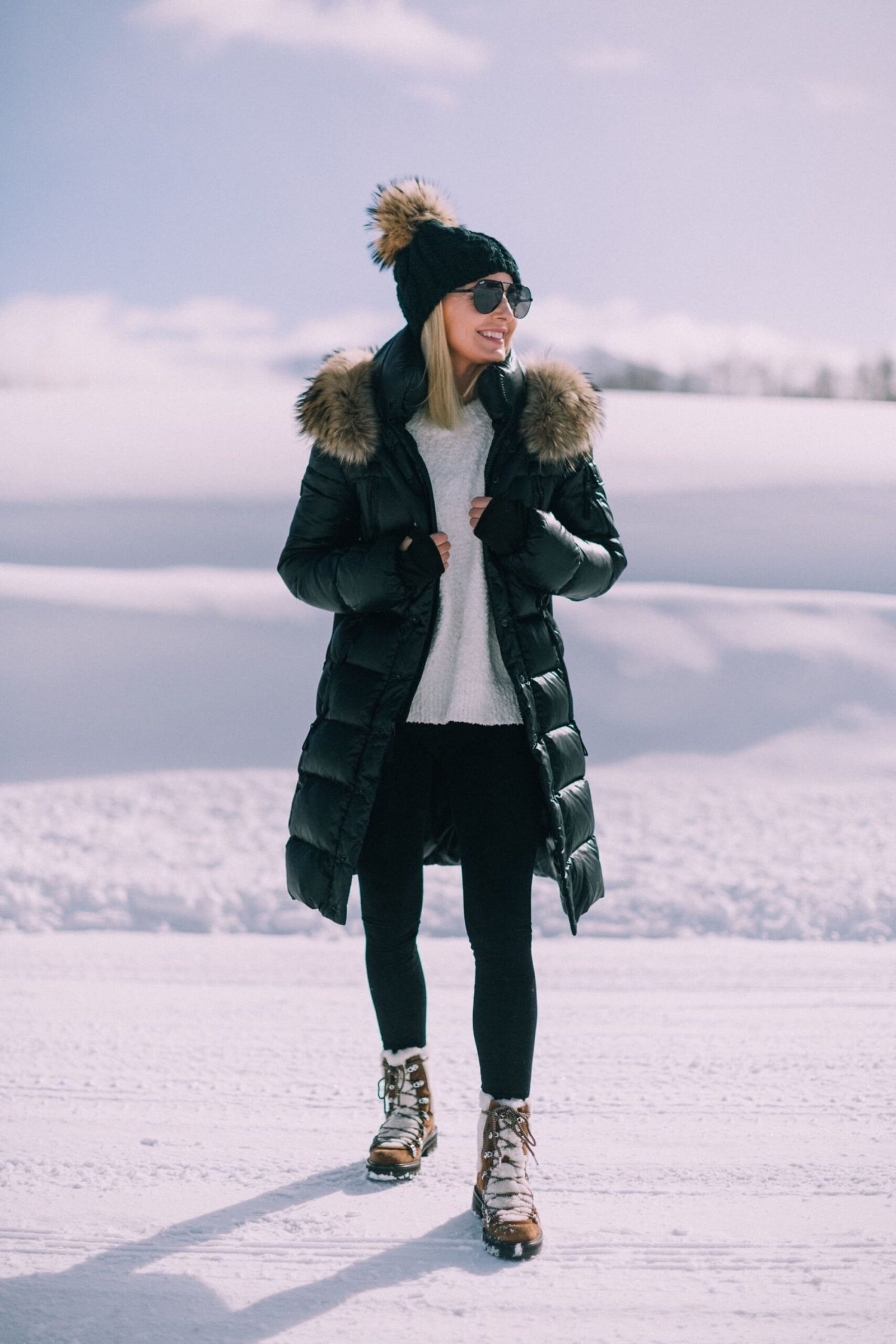 Older woman in black puffer with faux fur hood and pom beanie in snowy forest