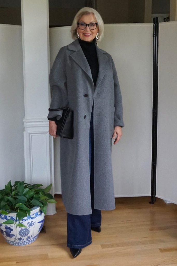 Winter outfit for women over 60: gray long coat, black turtleneck, jeans, indoors with plant