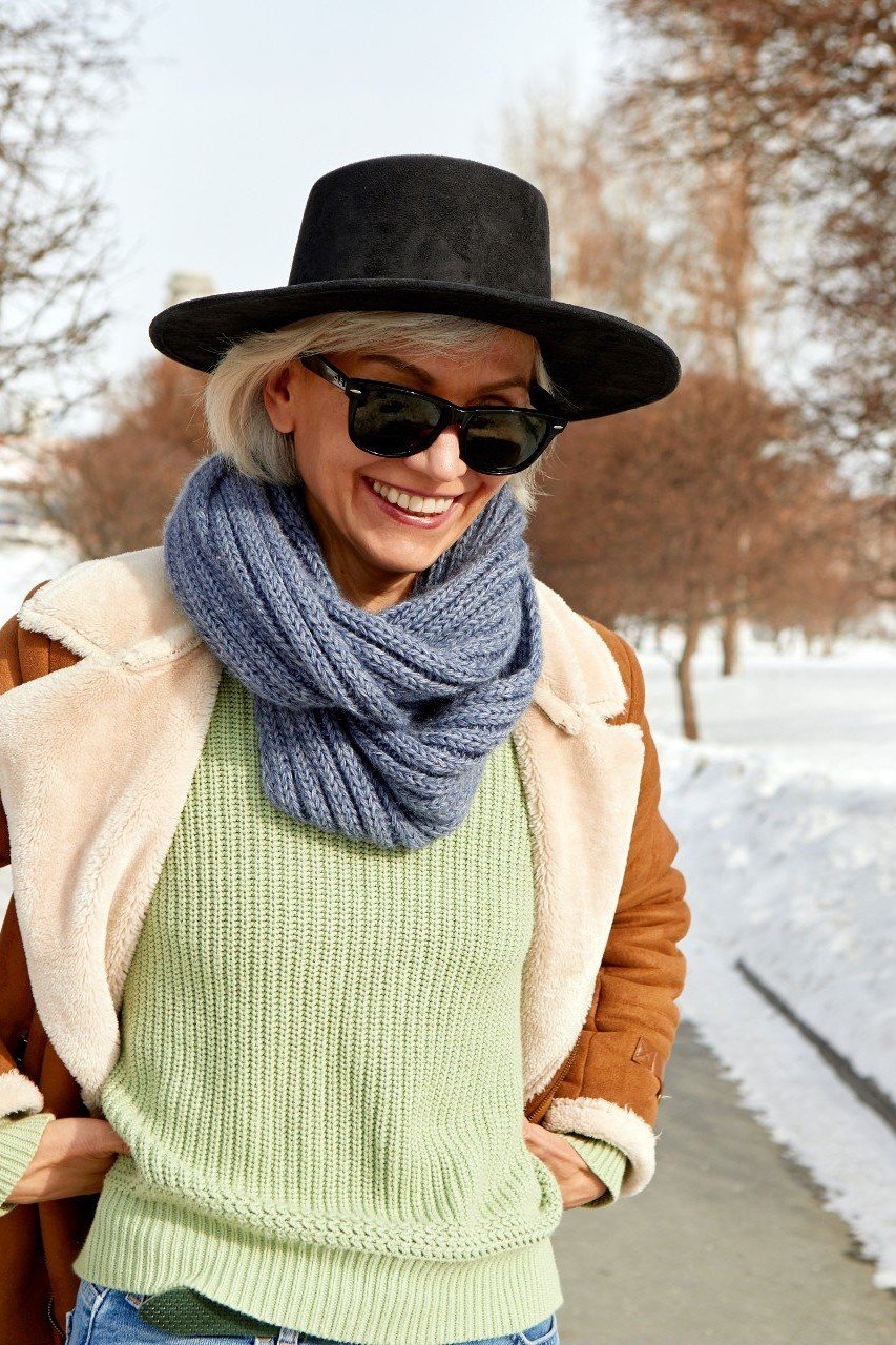 Winter outfit for women over 60: tan suede jacket, green sweater, blue scarf, black hat, snow
