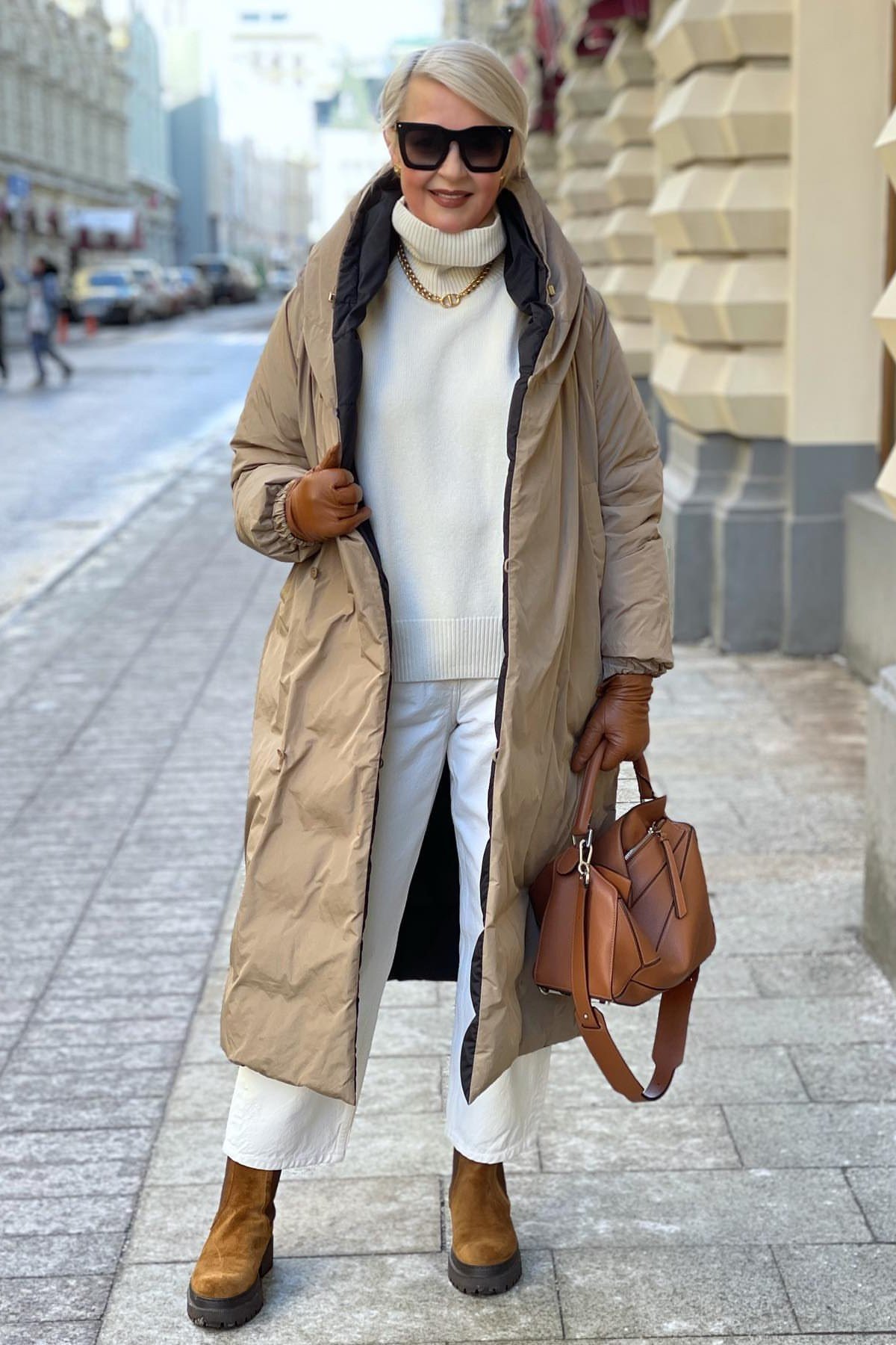 Winter outfit for women over 60: tan hooded parka, white turtleneck, white pants, tan boots, snow