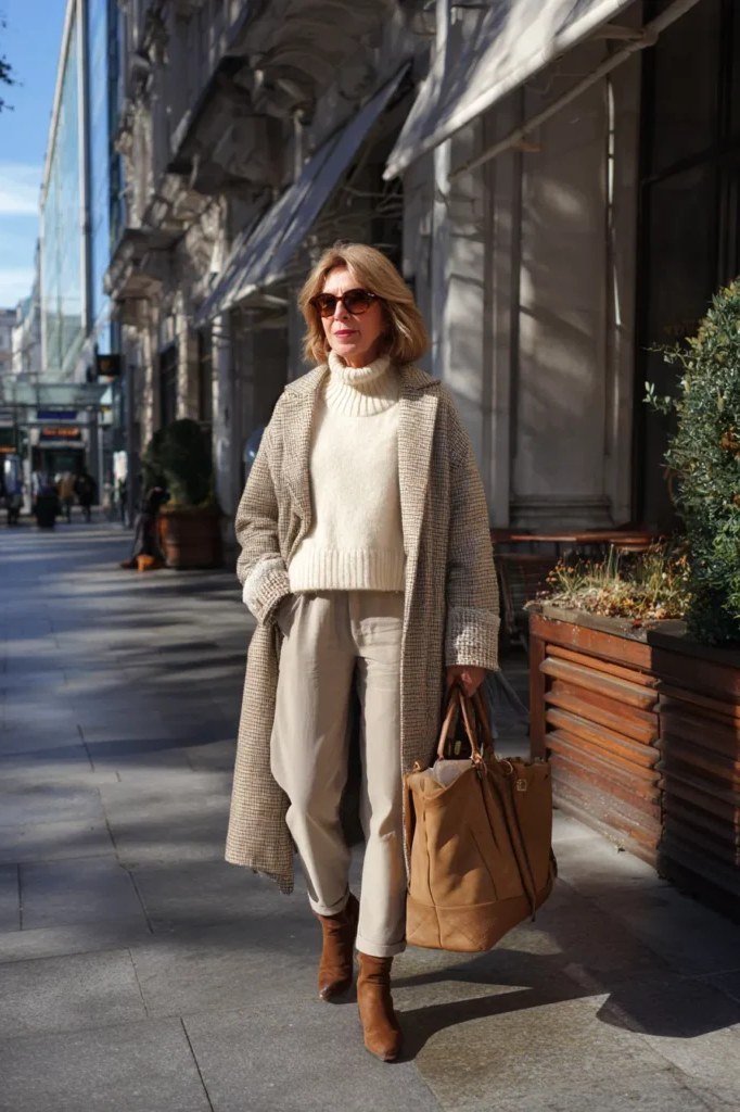 Winter outfit for women over 60: cream coat, cream sweater, tan handbag, tan boots, city