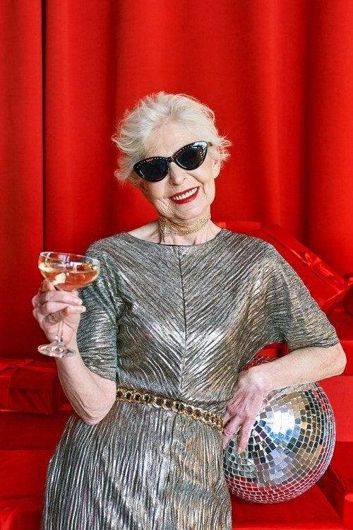 Smiling woman in silver pleated sequin dress with sunglasses holding champagne and disco ball, fun New Year's Eve outfit for women over 60