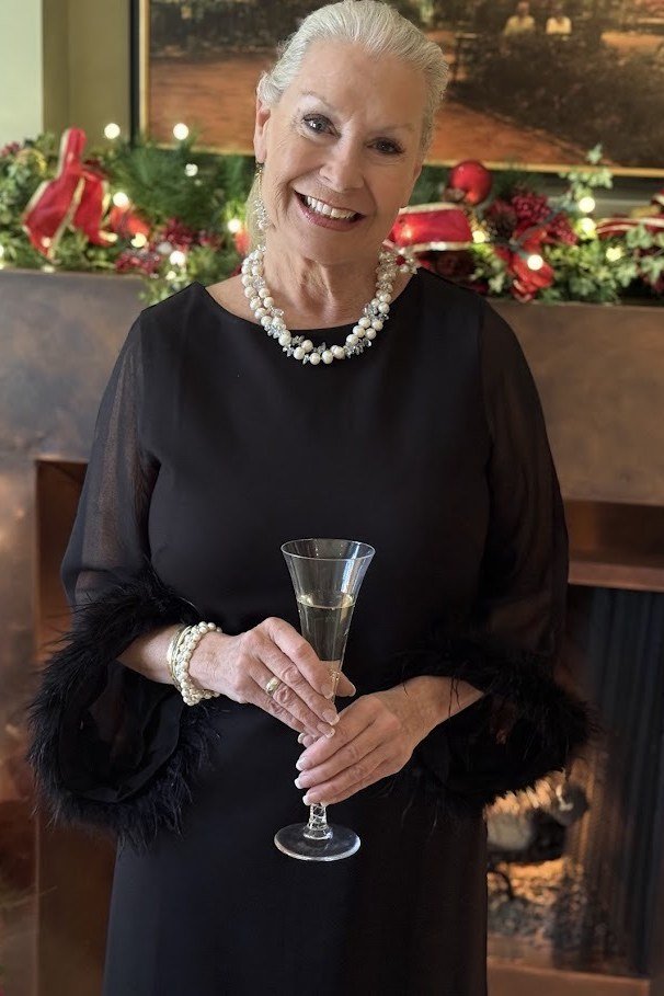 Smiling woman in black dress with feather-trimmed sleeves and pearl necklace holding champagne flute, chic New Year's Eve outfit for women over 60