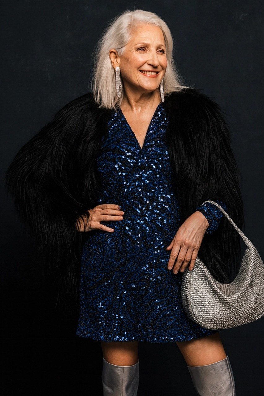 Woman in navy sequin dress with black faux-fur coat and silver handbag, full-length New Year's Eve outfit for women over 60