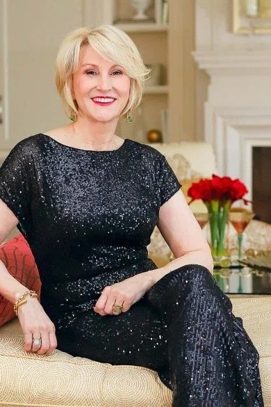Blonde woman in black sequin short-sleeve dress seated on sofa by fireplace, cozy New Year's Eve outfit for women over 60
