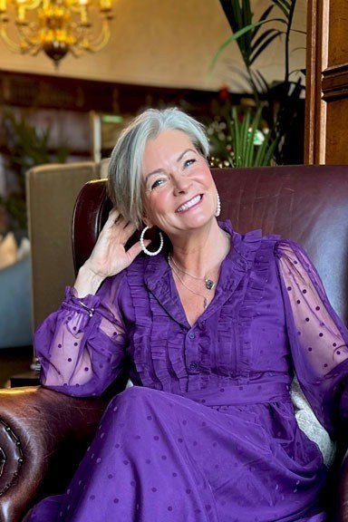 Gray-haired woman in purple dress with sheer sleeves seated holding wine glass, graceful New Year's Eve outfit for women over 60