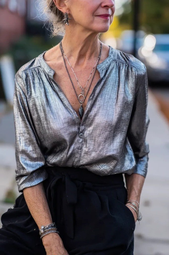 Gray-haired woman in metallic silver blouse with black pants and layered necklaces, polished New Year's Eve outfit for women over 60