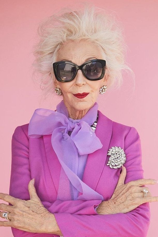 White-haired woman in fuchsia blazer with lilac bow blouse, brooch and sunglasses, colorful New Year's Eve blazer outfit for women over 60