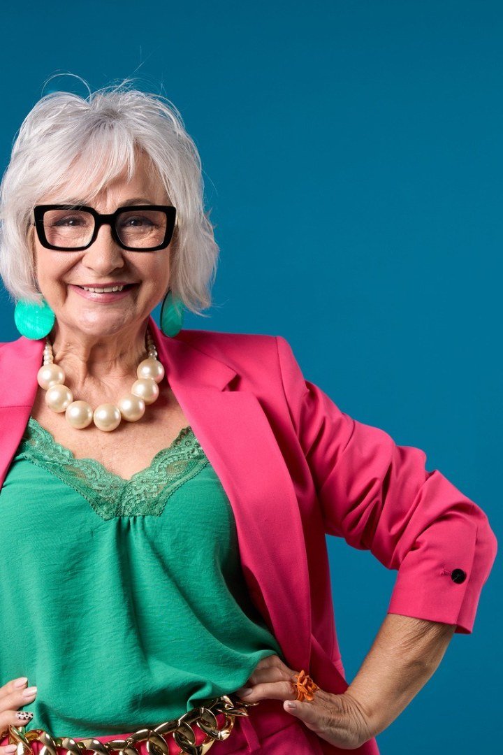 Woman in pink blazer with green lace top and pearl necklace against teal backdrop, pretty New Year's Eve blazer outfit for women over 60