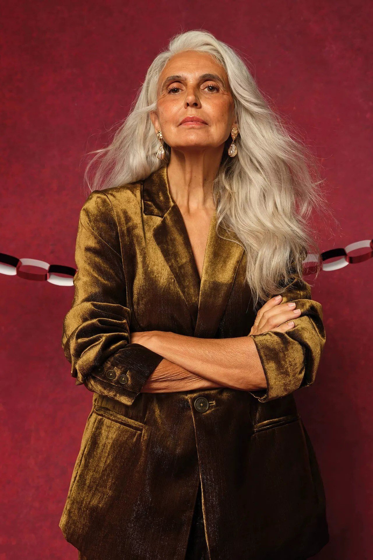 Woman with long silver hair in gold velvet blazer against red backdrop, luxe New Year's Eve blazer outfit for women over 60