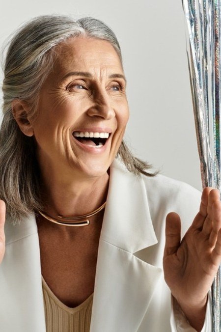 Laughing woman in white blazer by silver tinsel curtain, joyful New Year's Eve blazer outfit for women over 60