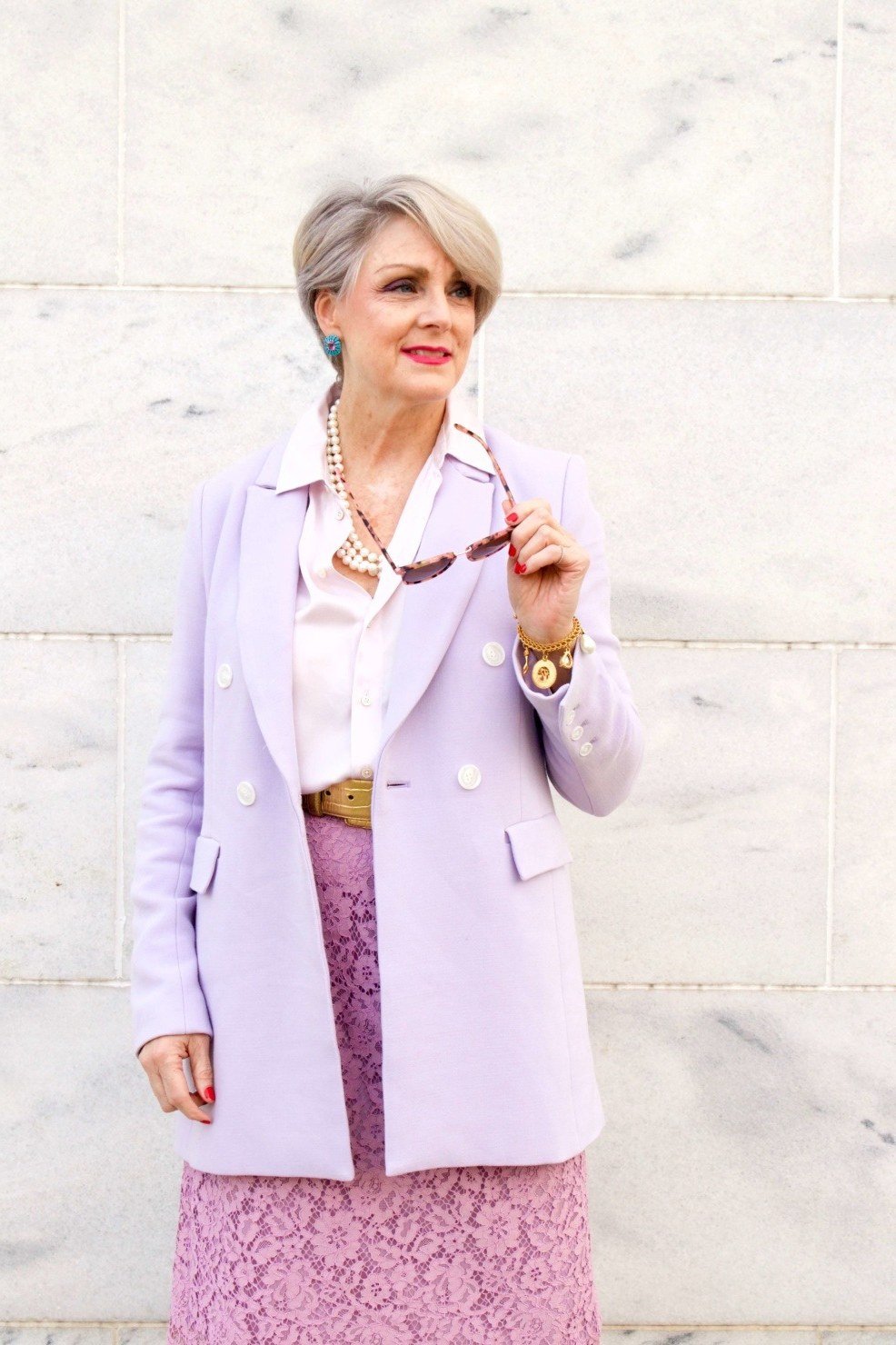 Woman in lavender blazer with pink lace skirt holding sunglasses, feminine New Year's Eve blazer outfit for women over 60