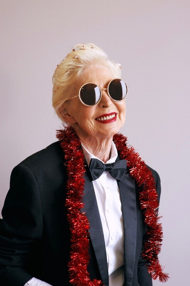 Smiling woman in black tuxedo with red tinsel boa and bow tie wearing sunglasses, fun New Year's Eve tuxedo outfit for women over 60