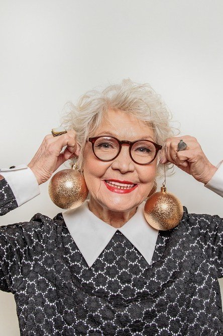 Smiling woman in patterned blazer holding up ornament earrings, festive New Year's Eve blazer outfit for women over 60